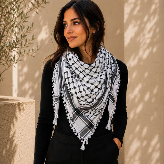 A woman wearing a black and white keffiyeh scarf with fringed edges, styled over a black long-sleeve top, standing against a sunlit beige stucco wall with leaf shadows.