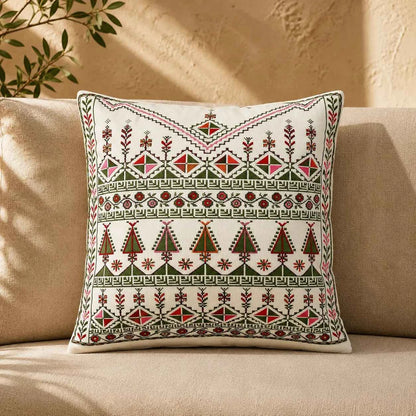 A square throw pillow with intricate geometric and floral embroidery in red, green, and black on an off-white background, resting on a beige sofa.