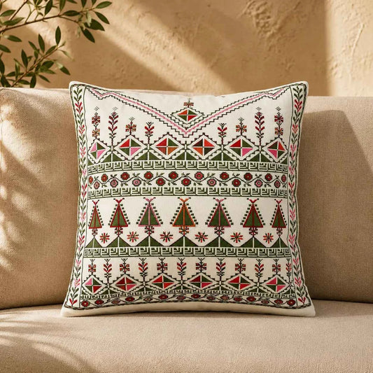 A square throw pillow with intricate geometric and floral embroidery in red, green, and black on an off-white background, resting on a beige sofa.