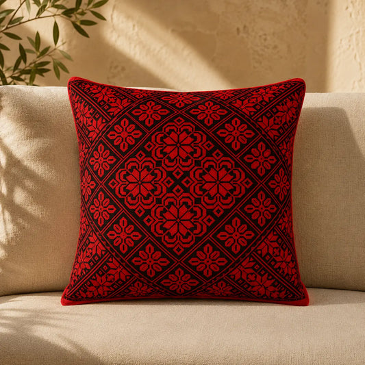 A square throw pillow with a red and black geometric floral pattern, resting on a beige fabric sofa in a sunlit room.